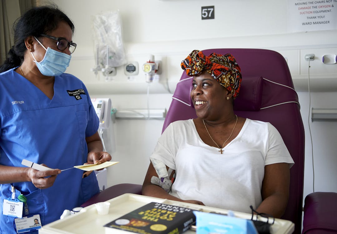 A clinical nurse specialist standing next to a hospital bed wearing her blue uniform and a blue face mask talking to a patient who’s sat in a purple chair with a drip in their arm