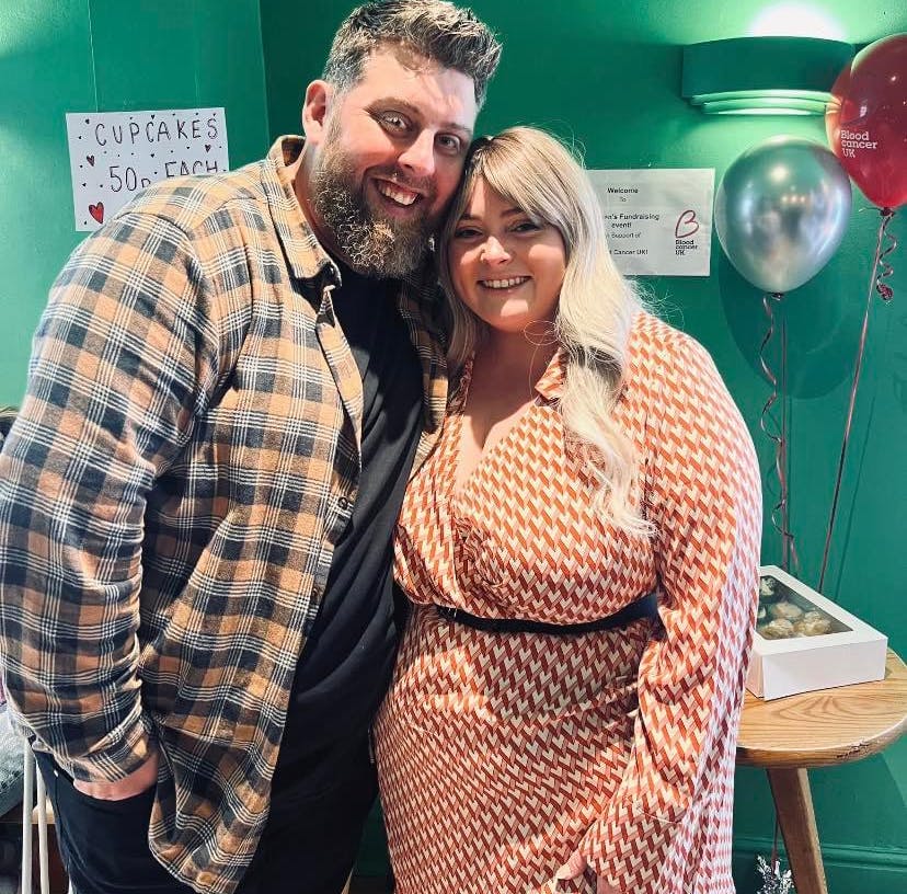 Bracken on the right wearing a red and white patterned dress with long blonde hair and her partner on the left with a grey beard and checkered shirt, standing with one arm around her and heads touching.