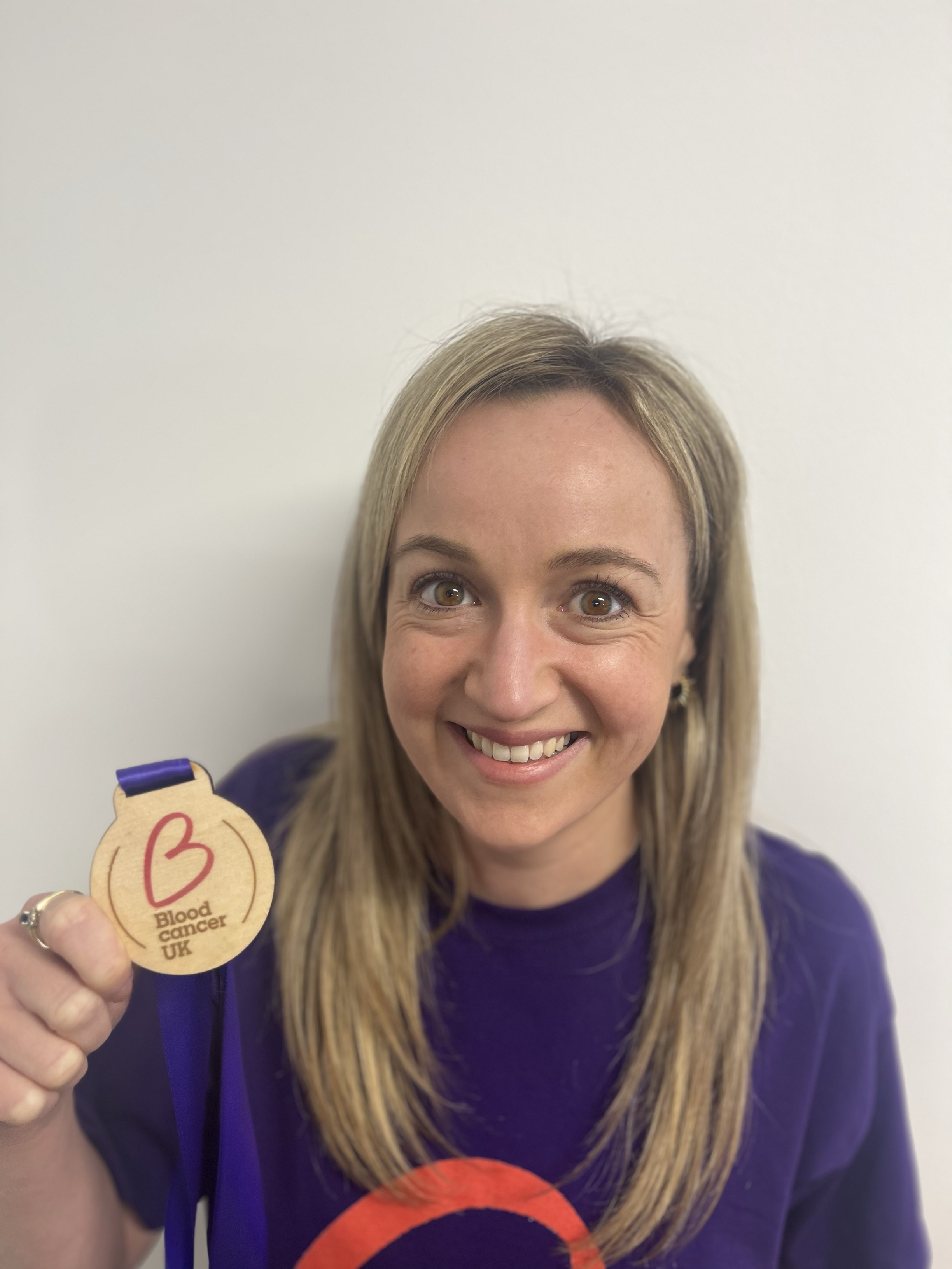 A woman with blonde hair holding a Blood Cancer UK medal up and smiling