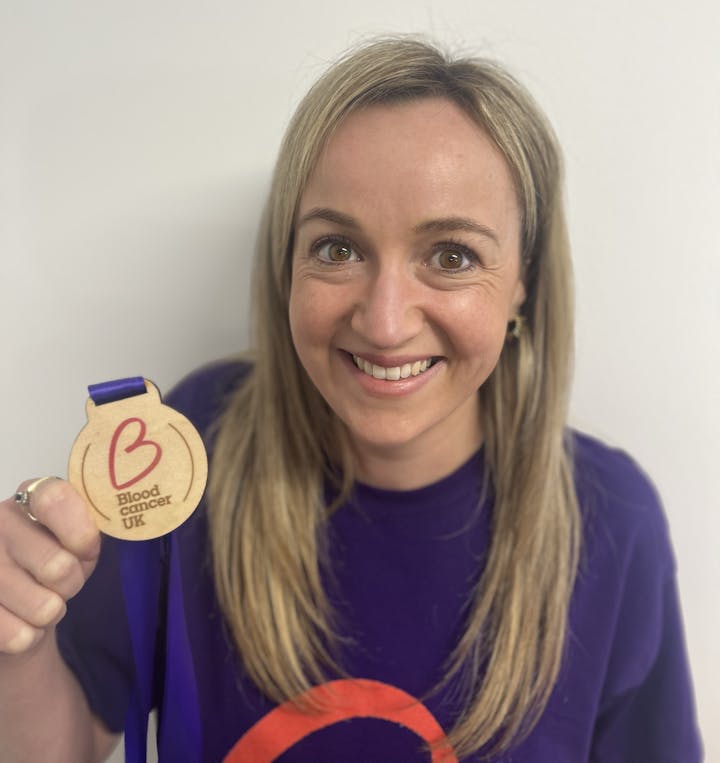 A woman with blonde hair holding a Blood Cancer UK medal up and smiling
