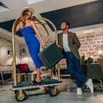 woman in blue dress holding a suitcase being pushed on a hotel luggage cart by man in smart casual blazer