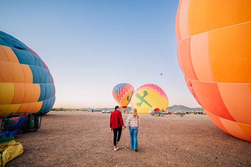 Man and woman holding hands while walking through the desert surrounded by hot air balloons