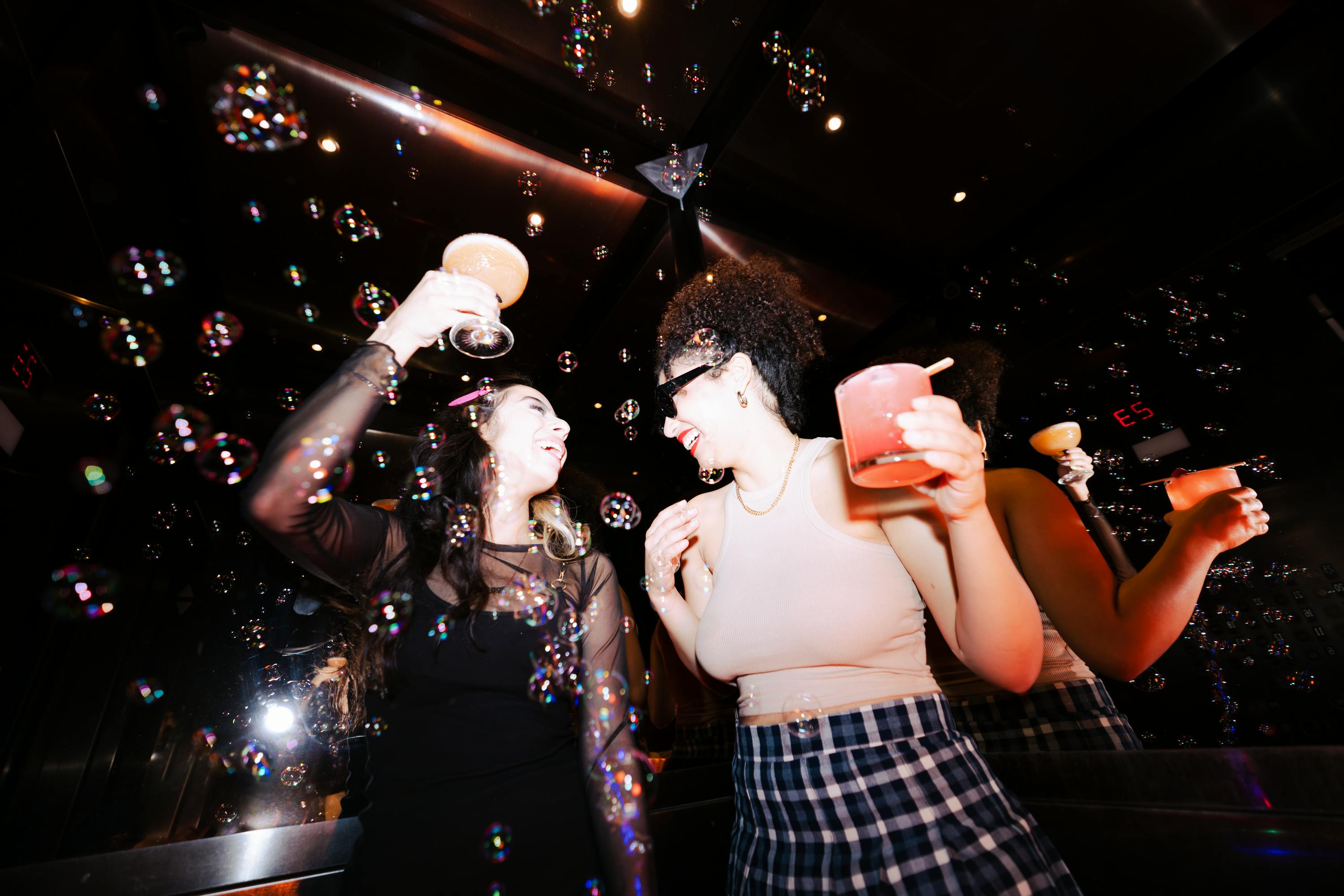 Two girls holding cocktails while dancing in a club surrounded by bubbles