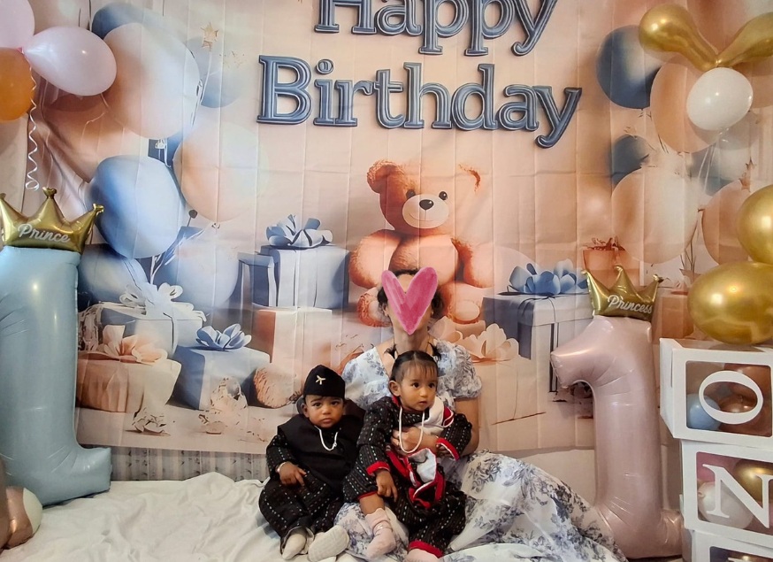 Annie sitting on the floor holding her dressed up twins in front of a 1st birthday photoshoot setup with decorations and gifts