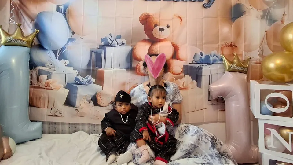 Annie sitting on the floor holding her dressed up twins in front of a 1st birthday photoshoot setup with decorations and gifts