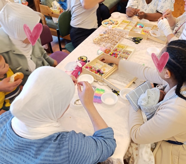 Women from different cultures sitting around a table doing beadwork. one of the woman holds a baby in yellow clothes