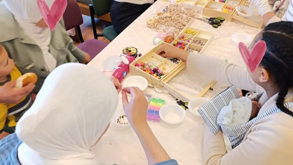 Women from different cultures sitting around a table doing beadwork. one of the woman holds a baby in yellow clothes