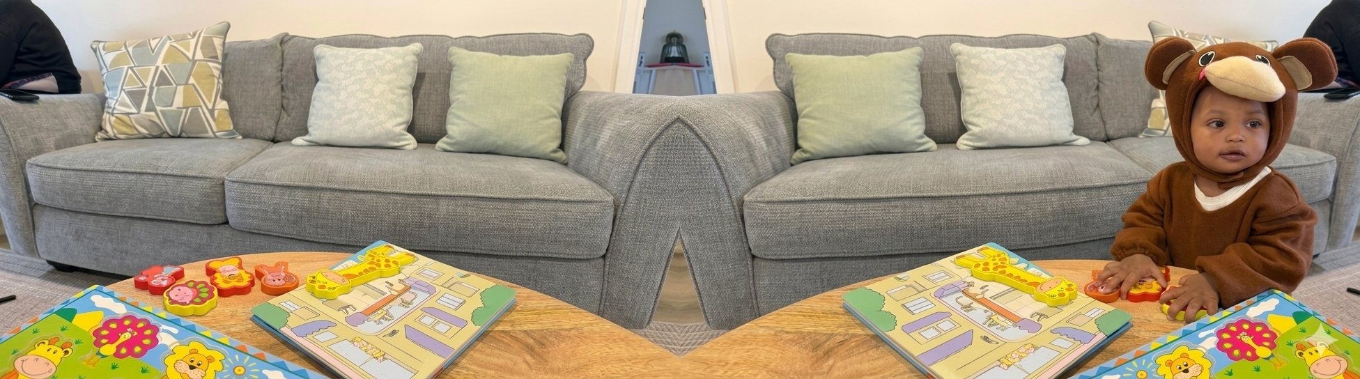 Baby Tarin, in een bruin apenpakje, kijkt naar boeken en speelgoed op een houten tafel, met daarachter een grijze bank met kussens.