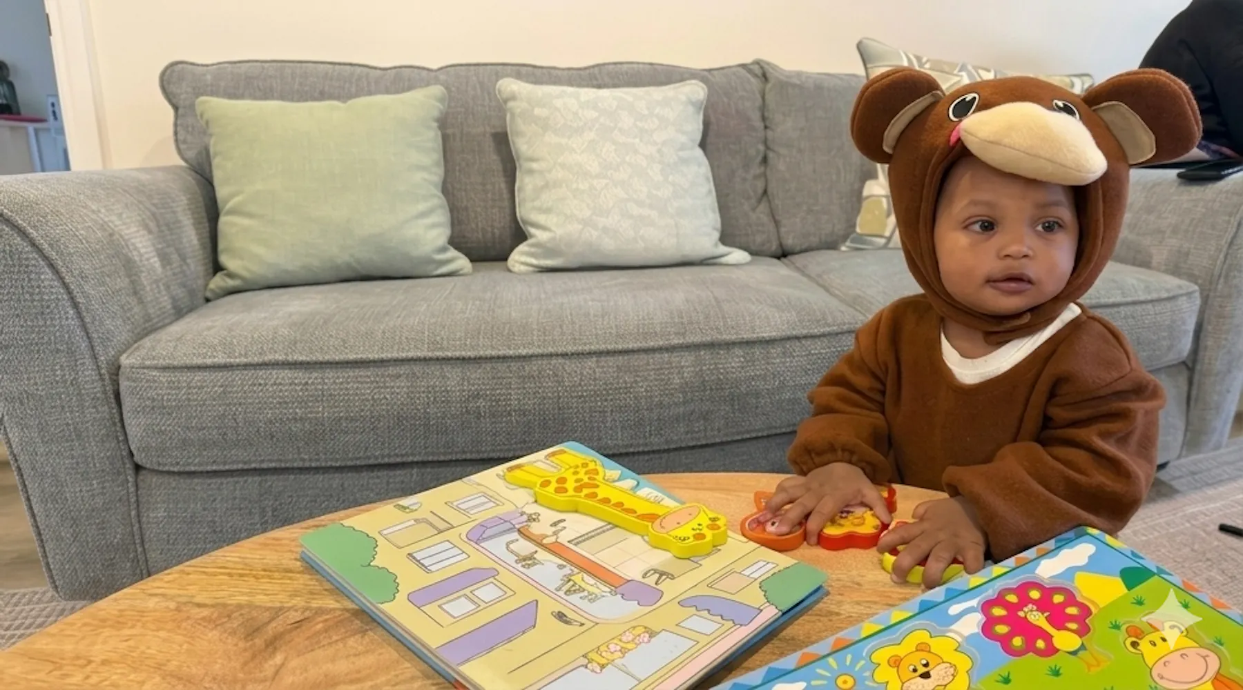 Baby Tarin, in een bruin apenpakje, kijkt naar boeken en speelgoed op een houten tafel, met daarachter een grijze bank met kussens.