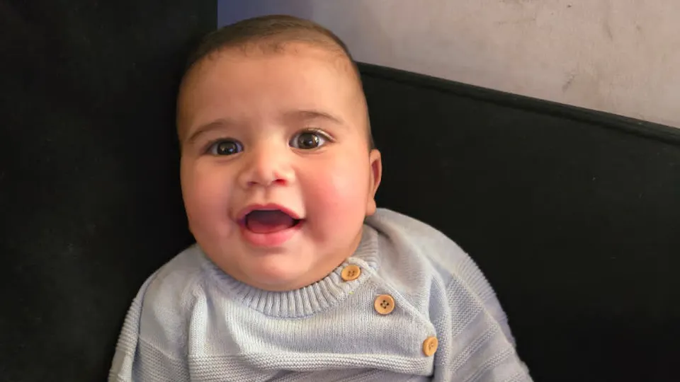 Noam met een brede glimlach in een blauwe raglan trui met knoopjes.