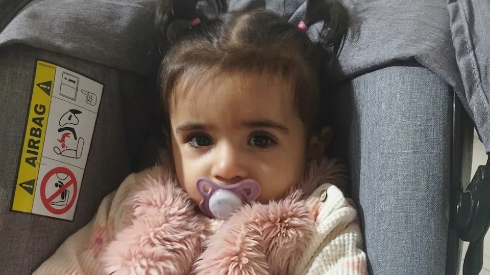 baby hallel in pink clothes, pacifier and a little pigtail in her dark hair, sitting in her car seat