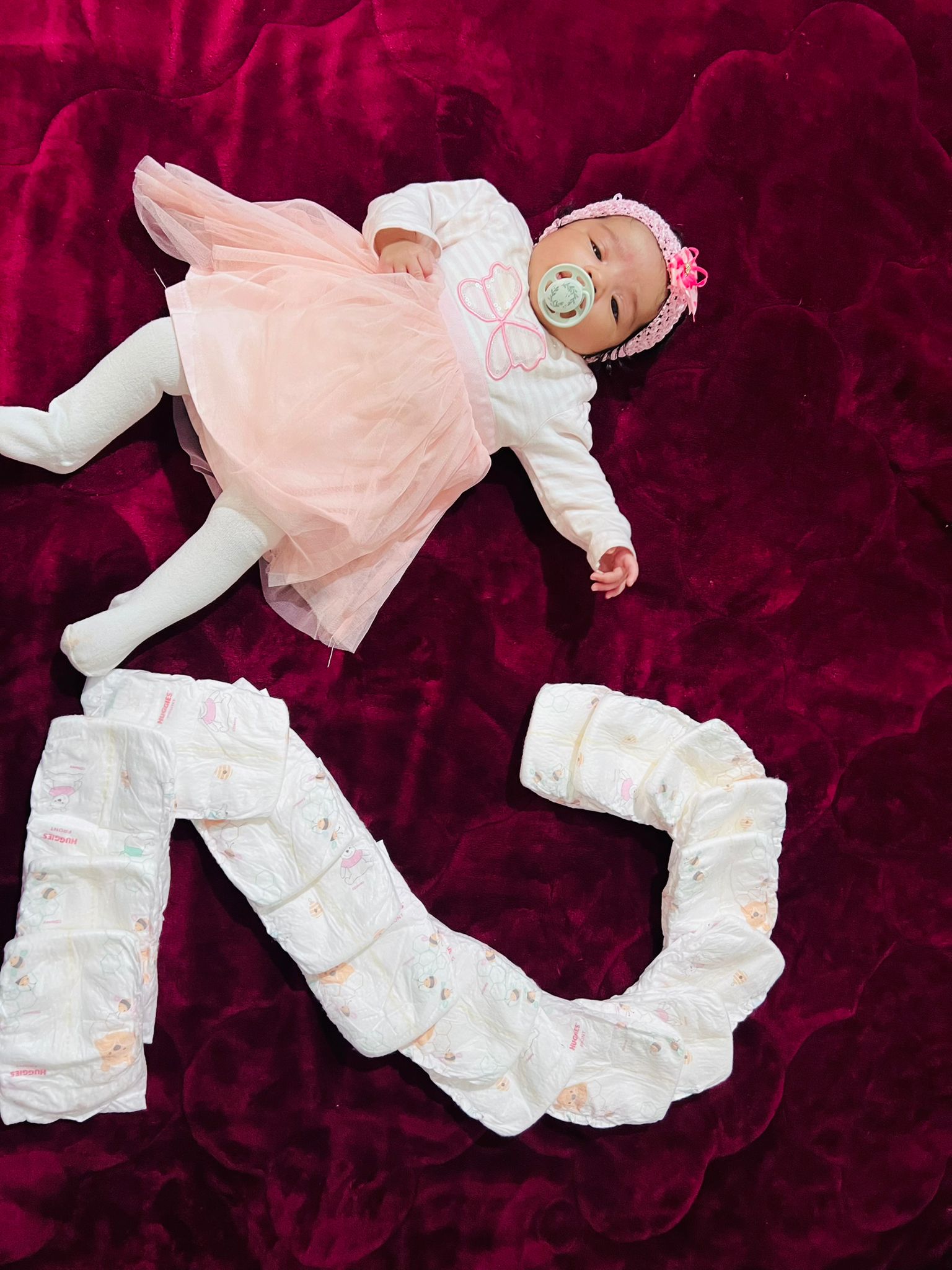 Baby Liana in pink and white dress next to a 2 built by diapers on a bordeaux colored blanket