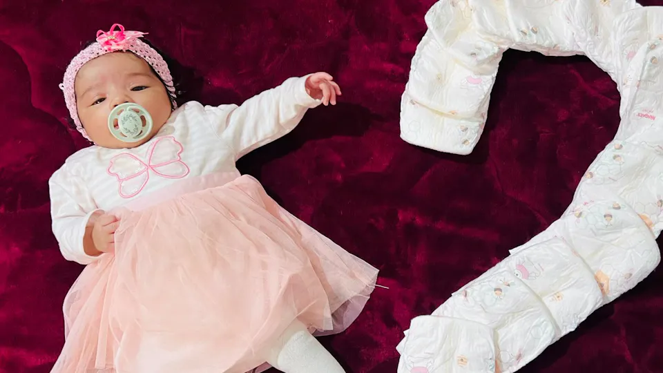 Baby Liana in pink and white dress next to a 2 built by diapers on a bordeaux colored blanket