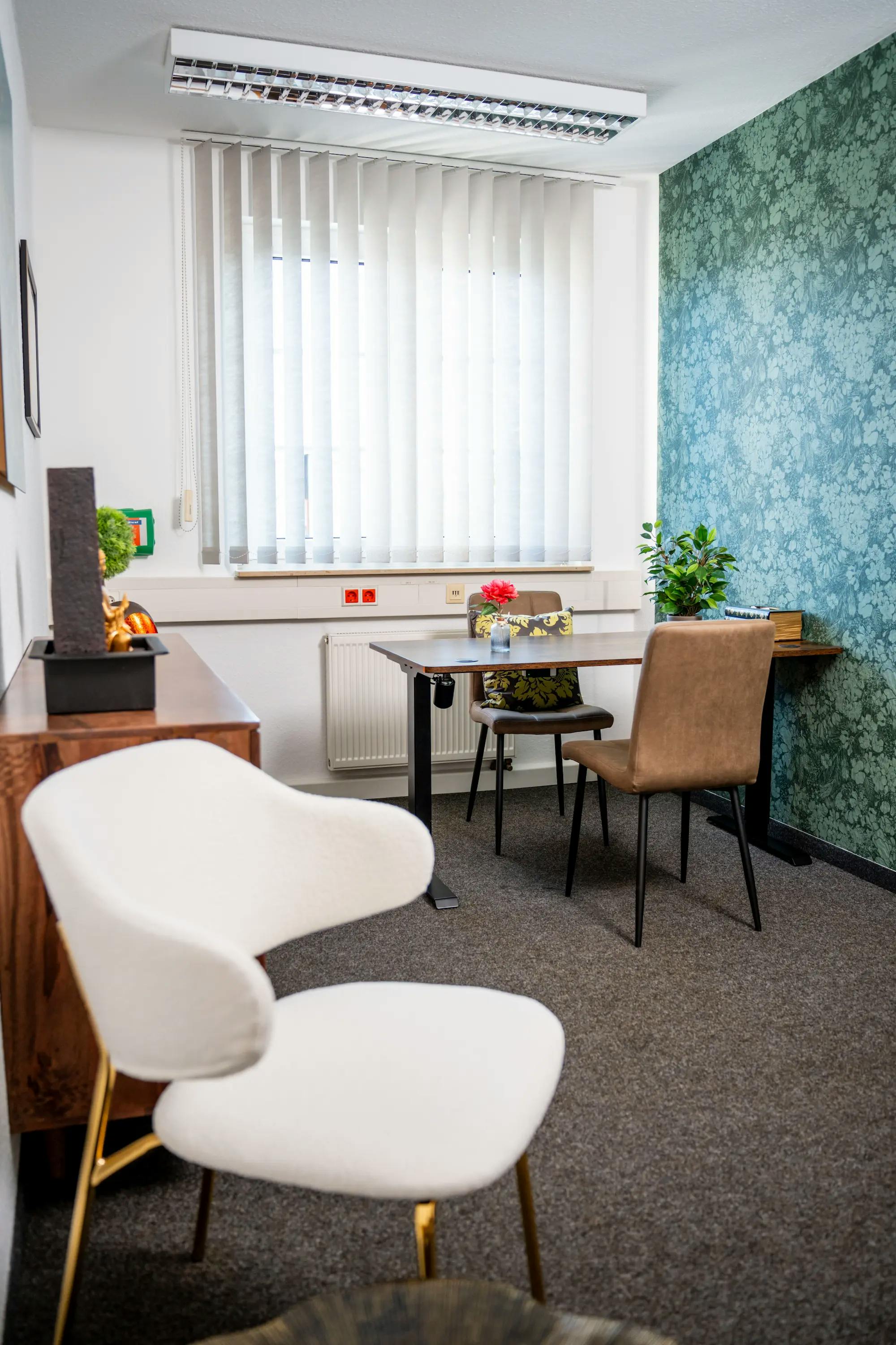 Helles Büro mit blauer, gemusterter Tapete. Im Vordergrund steht ein weißer Stuhl. Im Hintergrund befindet sich ein Tisch mit zwei Stühlen.