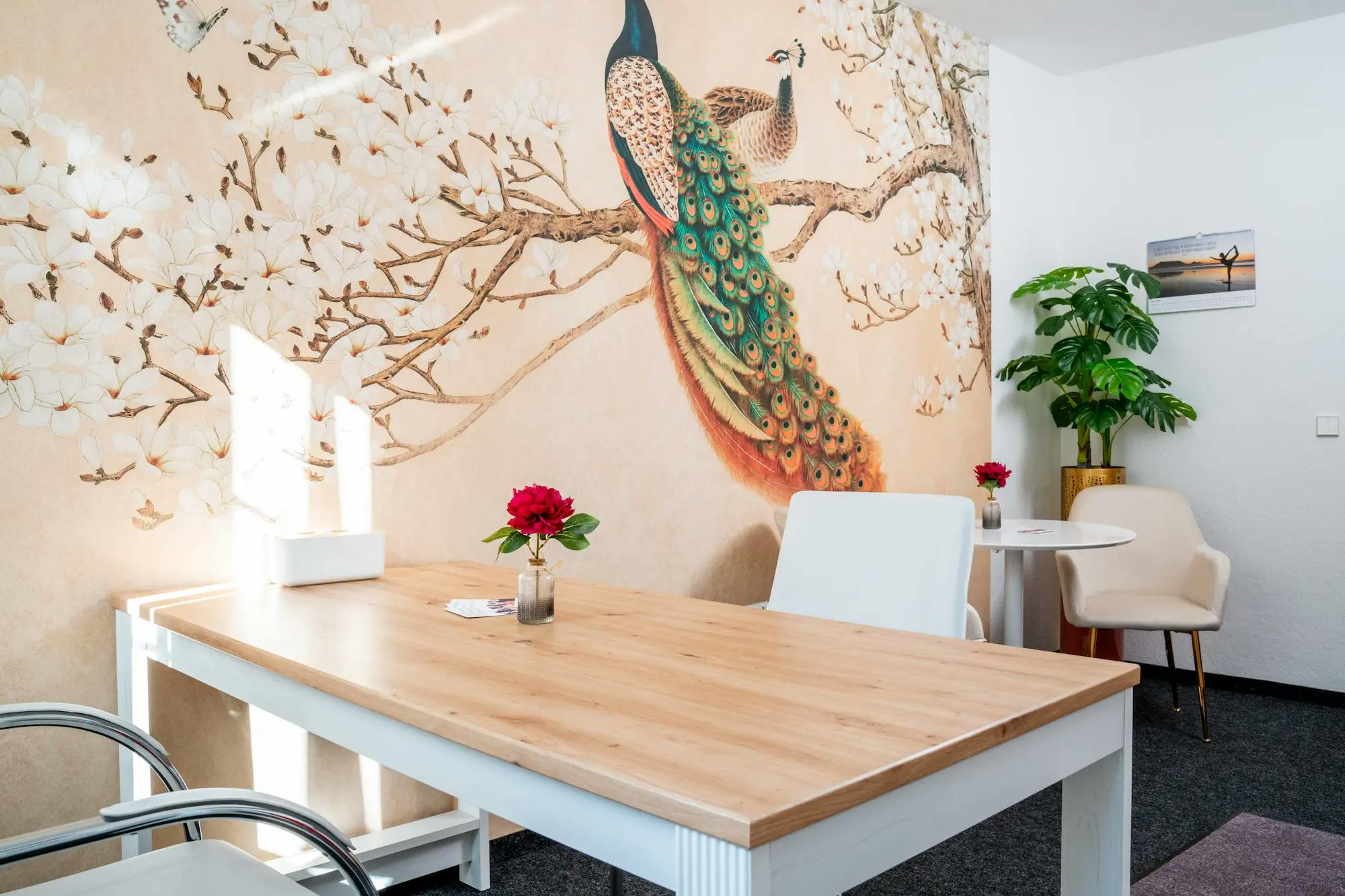 Weißer Tisch mit zwei weißen Stühlen vor hellbrauner Tapete mit einem Wandbild von einem Pfau auf einem Ast