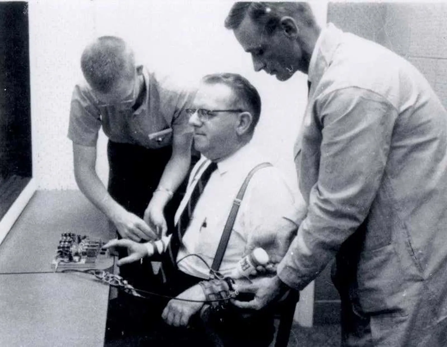 Zwart-witfoto van het Milgram-experiment: een zittende man krijgt elektroden op zijn polsen bevestigd door twee mannen, van wie één een witte laboratoriumjas draagt.