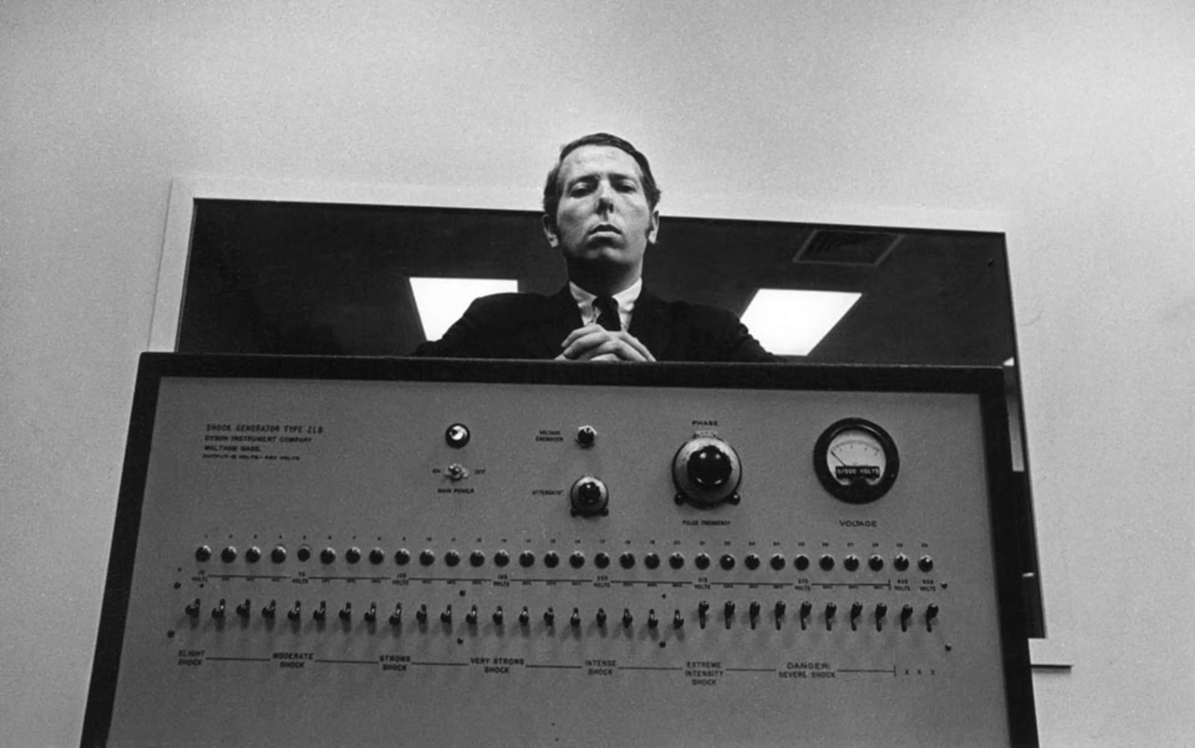 Een zwart-witfoto van psycholoog Stanley Milgram, die streng in de camera kijkt terwijl hij achter zijn beruchte "shock generator" staat. Het apparaat op de voorgrond bevat een lange rij van 30 schakelaars met tekstuele labels die oplopen in intensiteit, variërend van "Slight Shock" tot "XXX". Milgram draagt een donker pak en kijkt neer op de kijker vanuit een laag camerastandpunt, wat een autoritaire indruk wekt.
