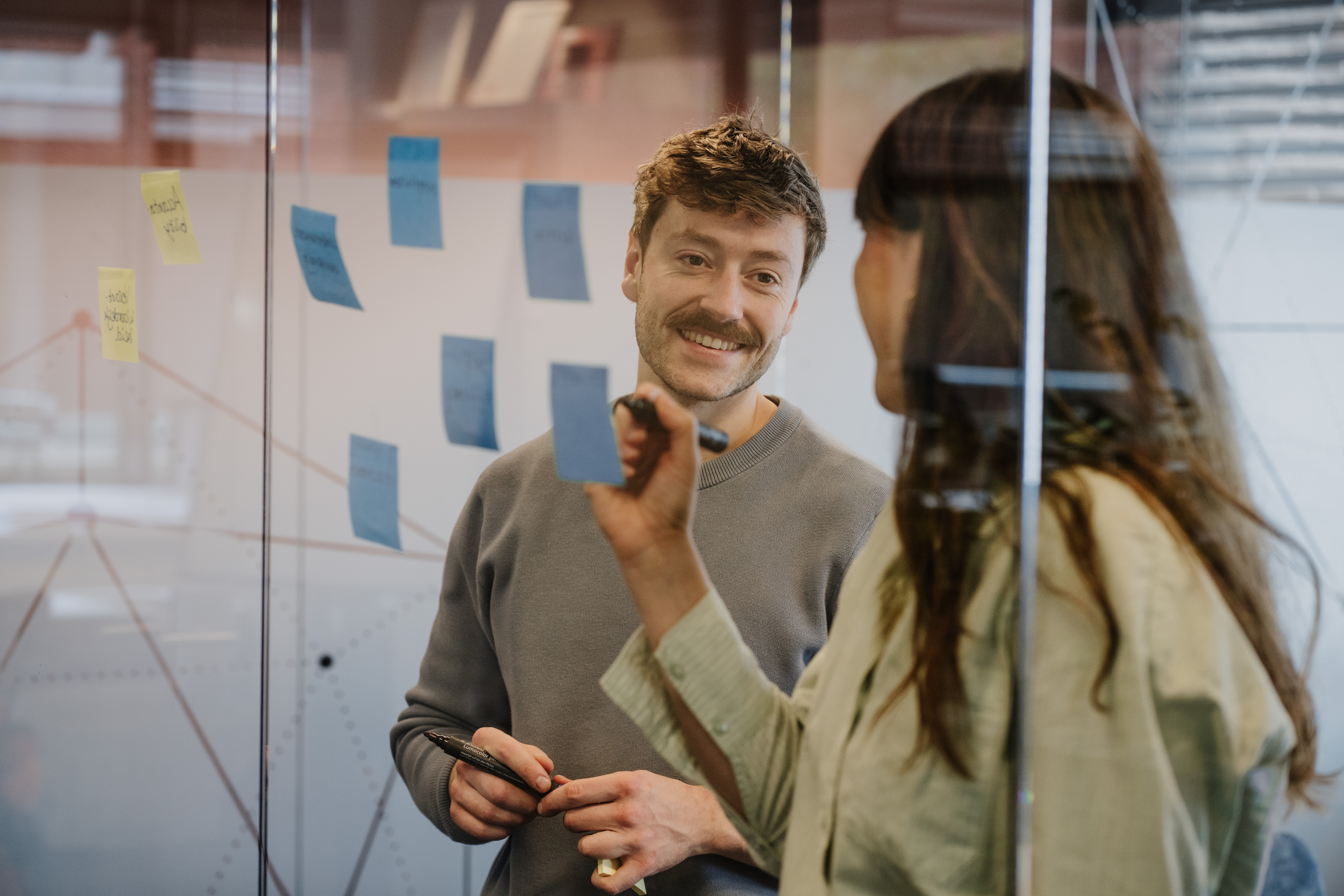 Een man en vrouw staan voor een glazen wand waar ze post-its op plakken.