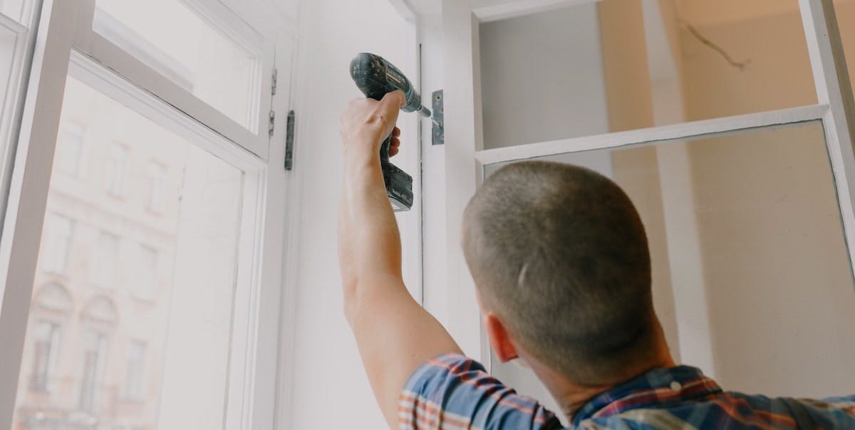 A young man with a hand drill secures window shutters as part of annual home maintenance on a rental home. 