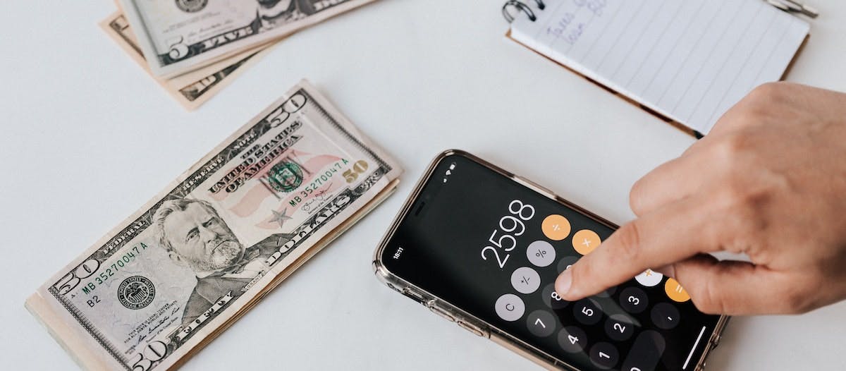 An image of a hand using an iphone calculator, depicting a person assessing if they can live off of rental property income, with American banknotes and a notepad beside them