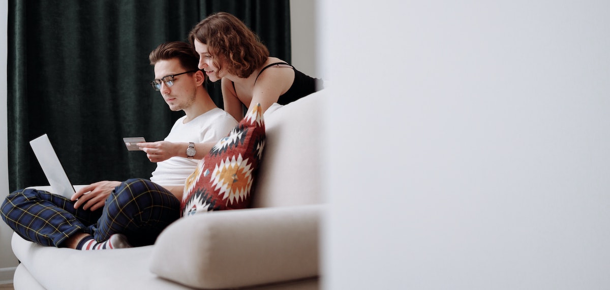 A young adult male sits at his laptop on the couch, while his female partner shows him a credit card, paying rent for their rental home securely online.