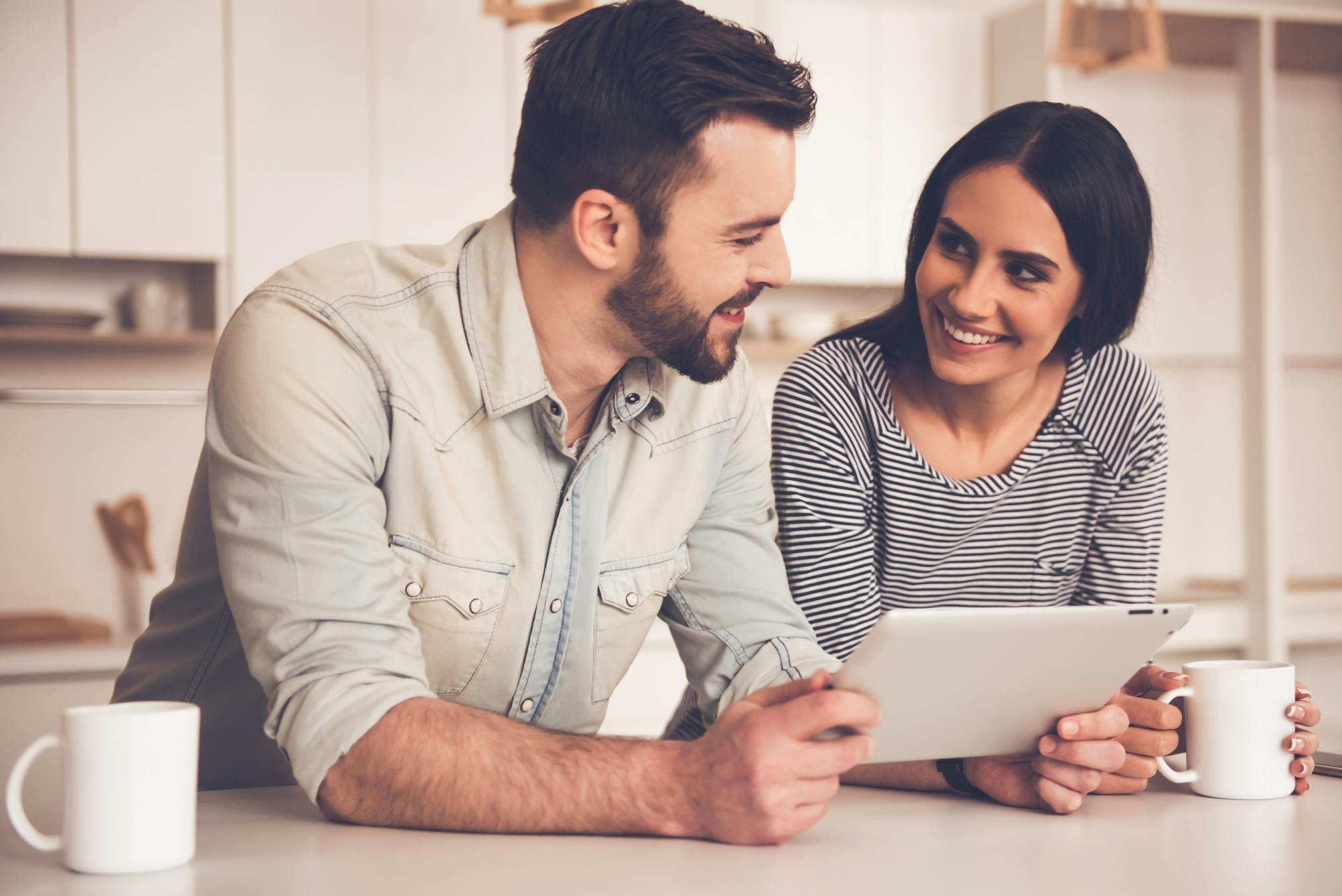 A young smiling couple hold a tablet device in the kitchen, with coffee. 