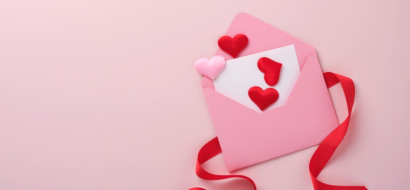 A pink envelope lays on a red ribbon, with a letter and love hearts spilling out, on a pink background. 