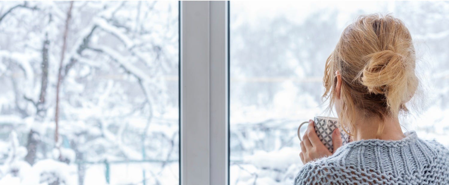 A woman holds a cup of tea while looking out the window at a snow-covered yard. Learn tips on winterizing your property to save time, money and headaches from the damage the cold and storms can create. 