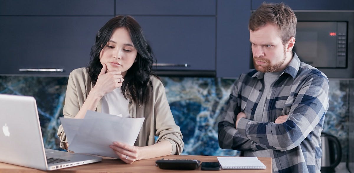 A couple stand in a kitchen with a laptop and calculator, looking concerned over unplanned fees and charges. Uncover property management fees that are costing landlords a fortune.