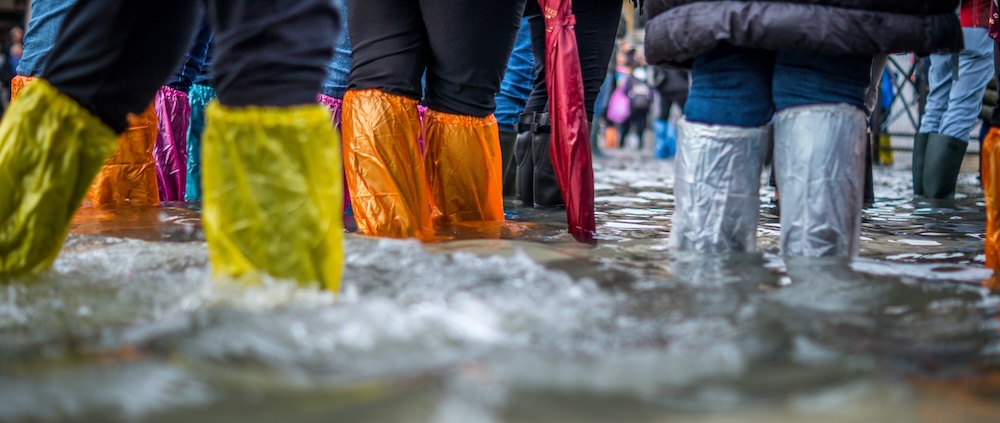 A crowd of people stand in flood water wearing plastic shoe protectors in yellow, orange pink and white