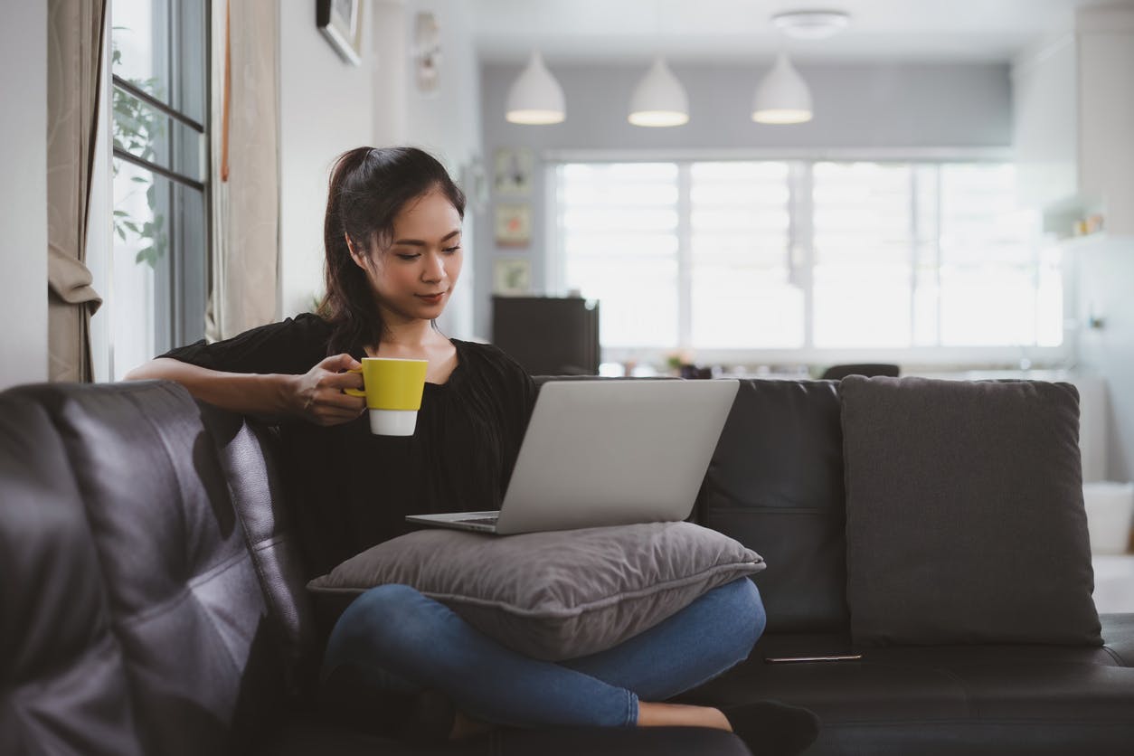 Young woman drinking coffee on the couch and working on her laptop, managing a rental from interstate