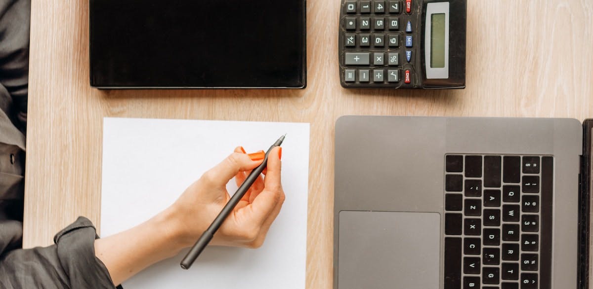 An image of a female hand with a pen, laptop and calculator, ready to complete a cap rate formula. Capitalization rates compare and estimate the future ROI of real estate investment properties.