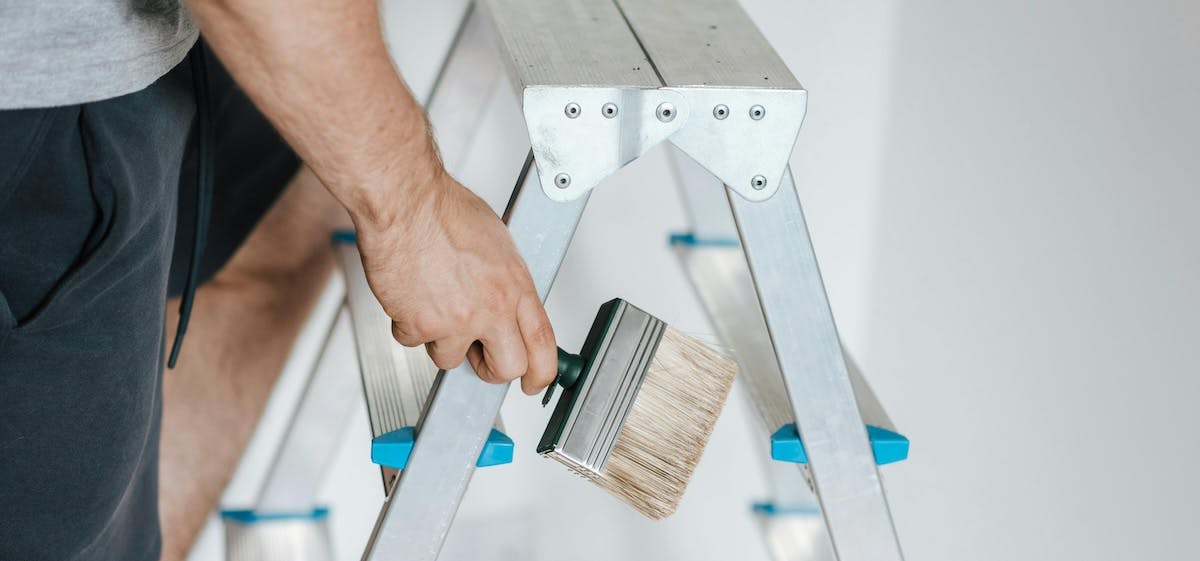 A cropped close up shot of a man holding a paintbrush on a step ladder