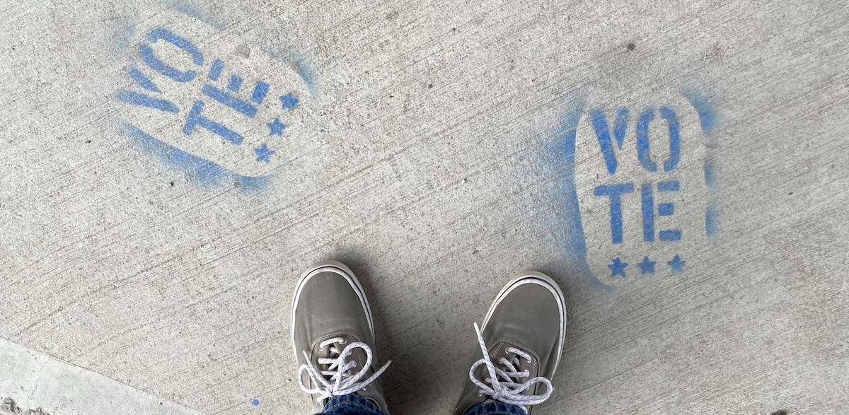 A pair of grey and white sneakers stands on concrete with "VOTE" spray painted on the ground