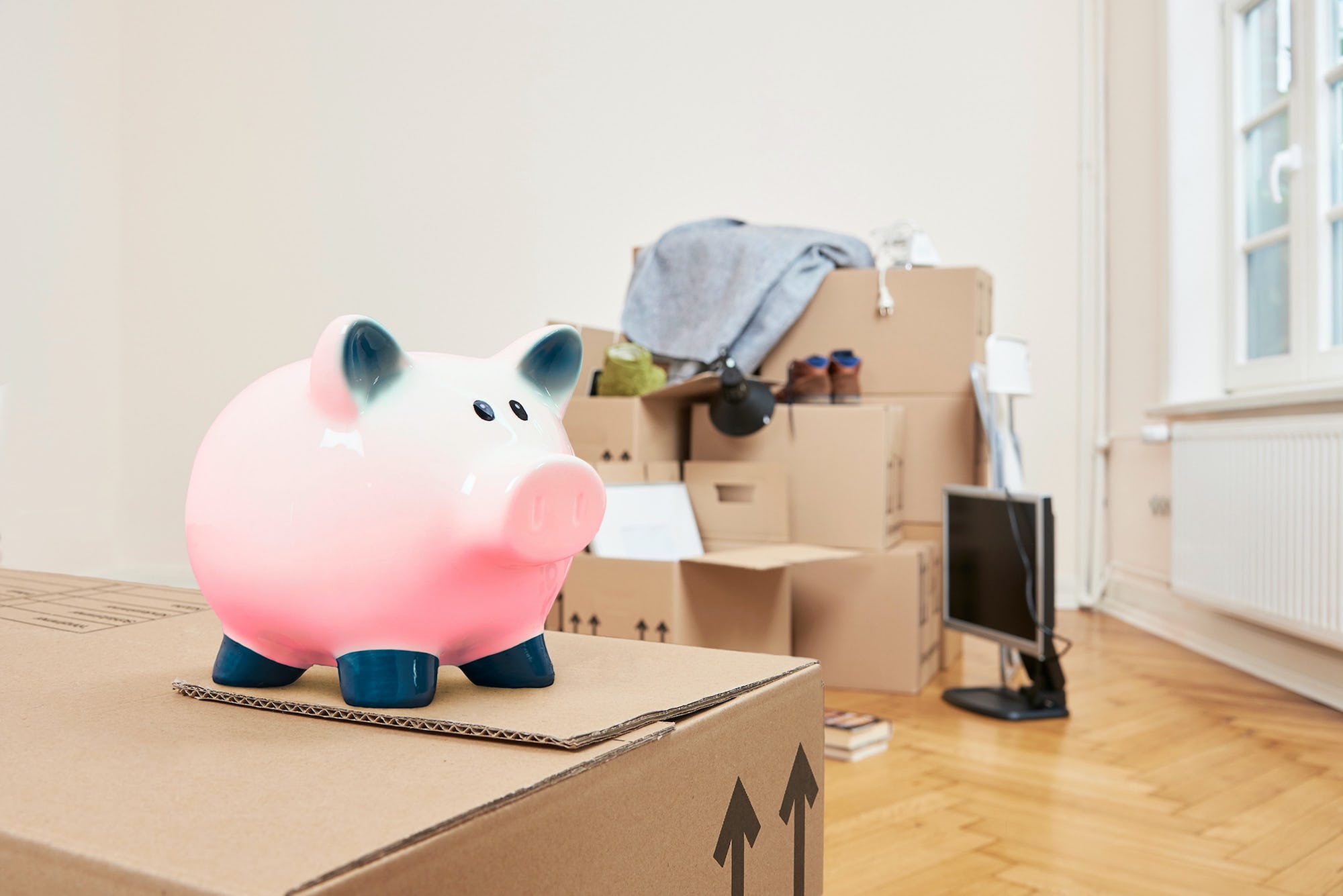 Moving boxes to relocate when it's time to downsize a home, with a pig piggy bank sitting on the moving box with household items in background