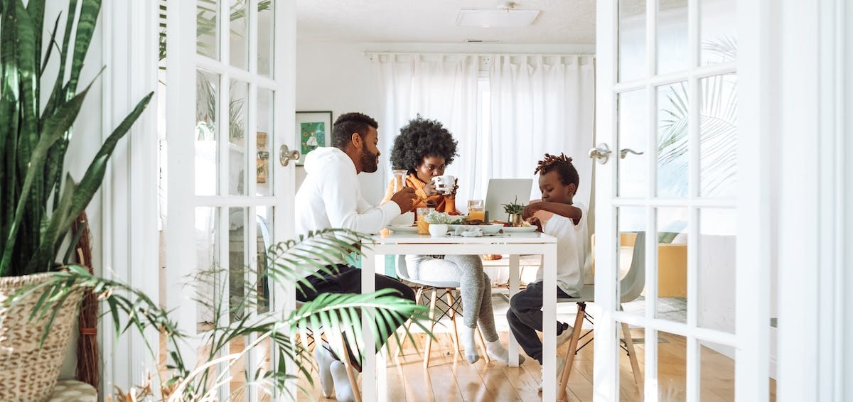 A view through open double-doors to show a family of three enjoying breakfast at a dining table with food and a laptop visible, depicting the sense of home and belonging