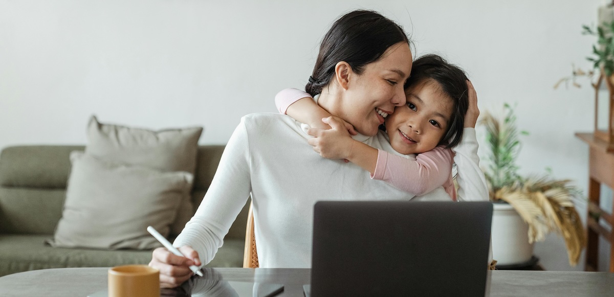 A smiling woman sits in front of a laptop while her young daughter gives her a hug from behind