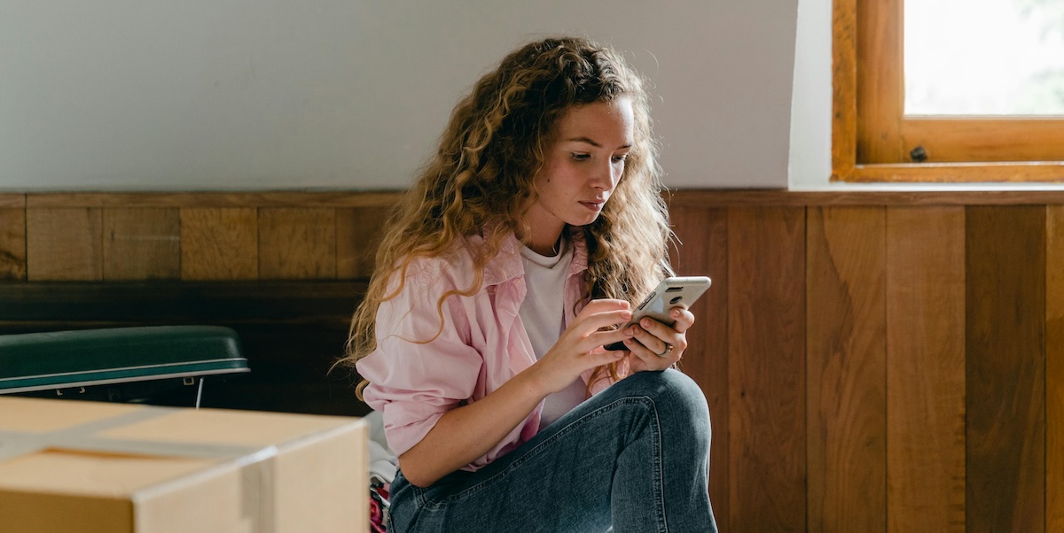 A young woman with long curly hair looks at her phone, surrounded by moving boxes