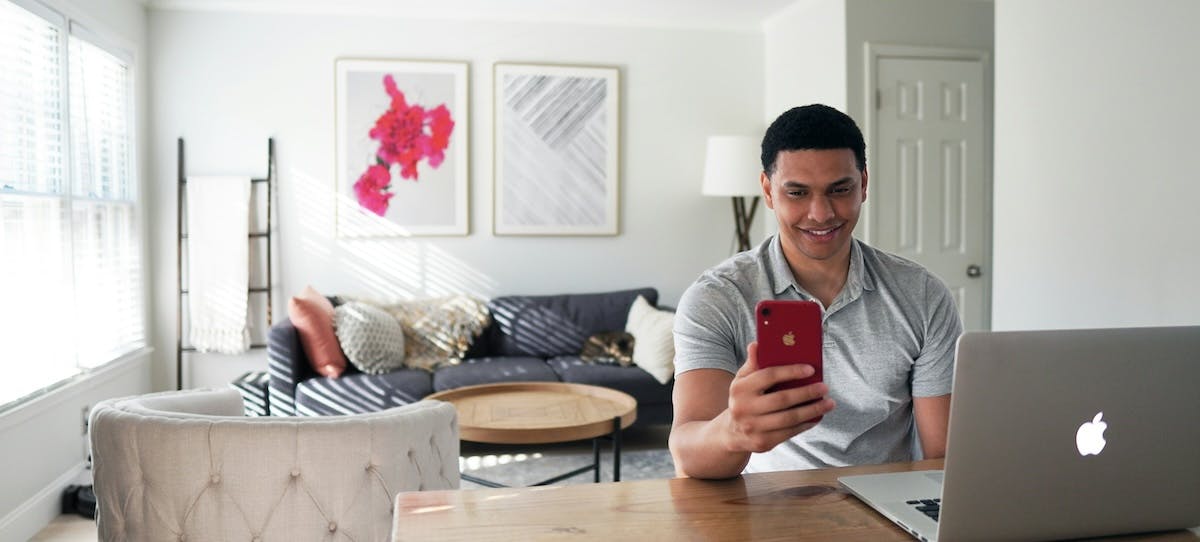 A young man sits at his laptop at home, with a red iphone in his hand, organizing his rental home finances online