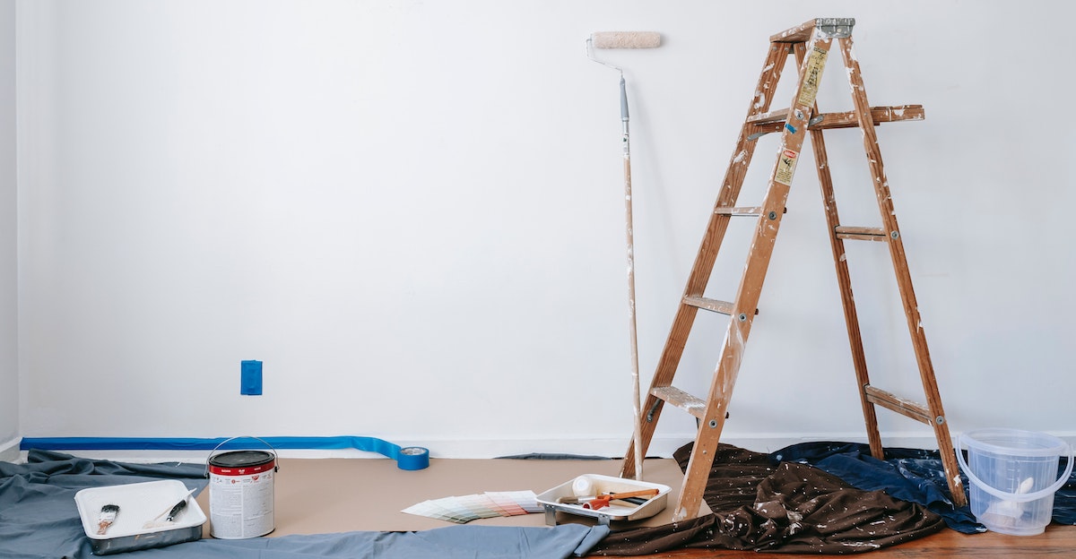 An image of home maintenance on a rental property, with a ladder, paint tin, roller, and paint brushes against a white wall.