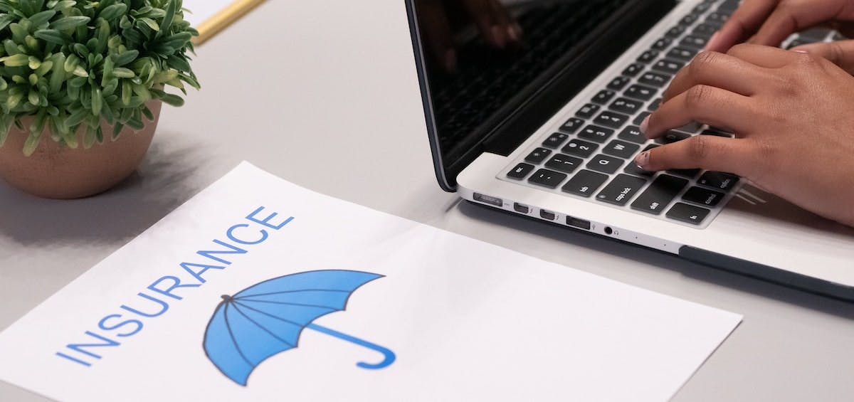A piece of paper shows the word "Insurance" above a blue umbrella, next to a laptop. Learn more about insurance for real estate investors. 
