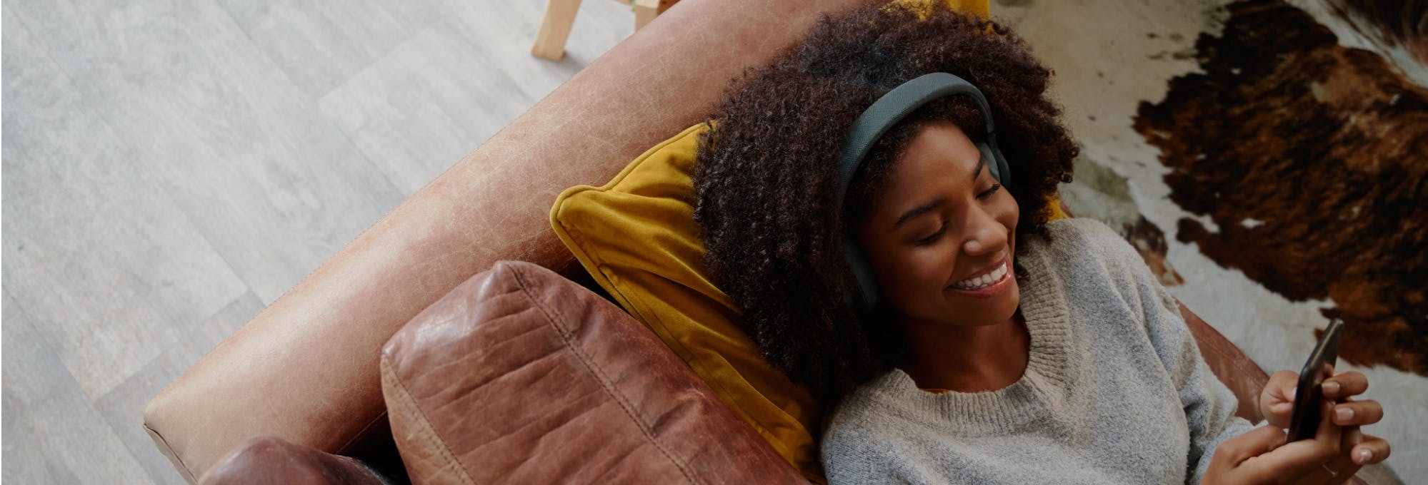A young woman of colour lays on a sofa looking at her phone, wearing headphones