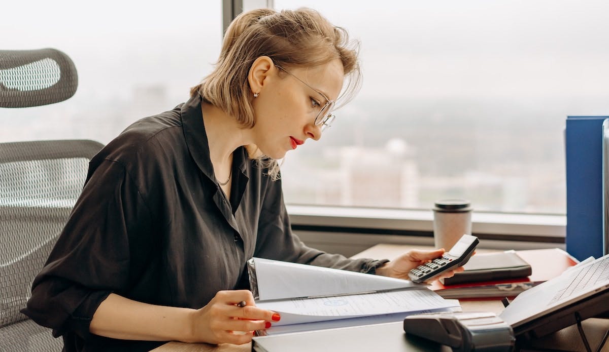 A female property accountant wearing glasses examines paperworks and figures on a calculator at an office with a city view in the background 