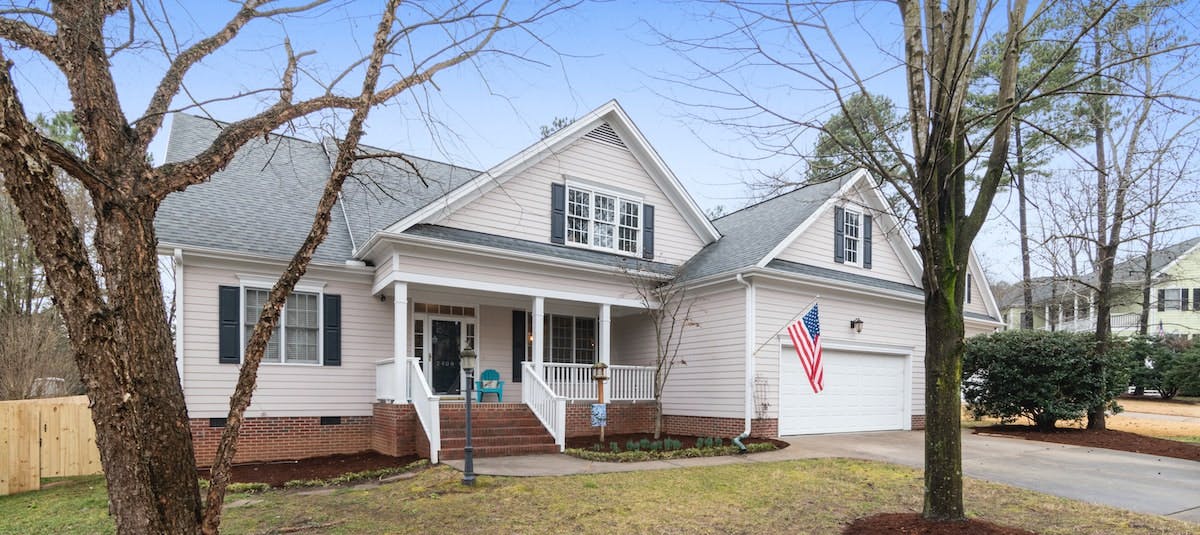 A photo of a white single-family home with an American flag and a grey roof, behind trees in the front yard. Learn about how US homeowners could be affected by 2022 ballot initiatives. 