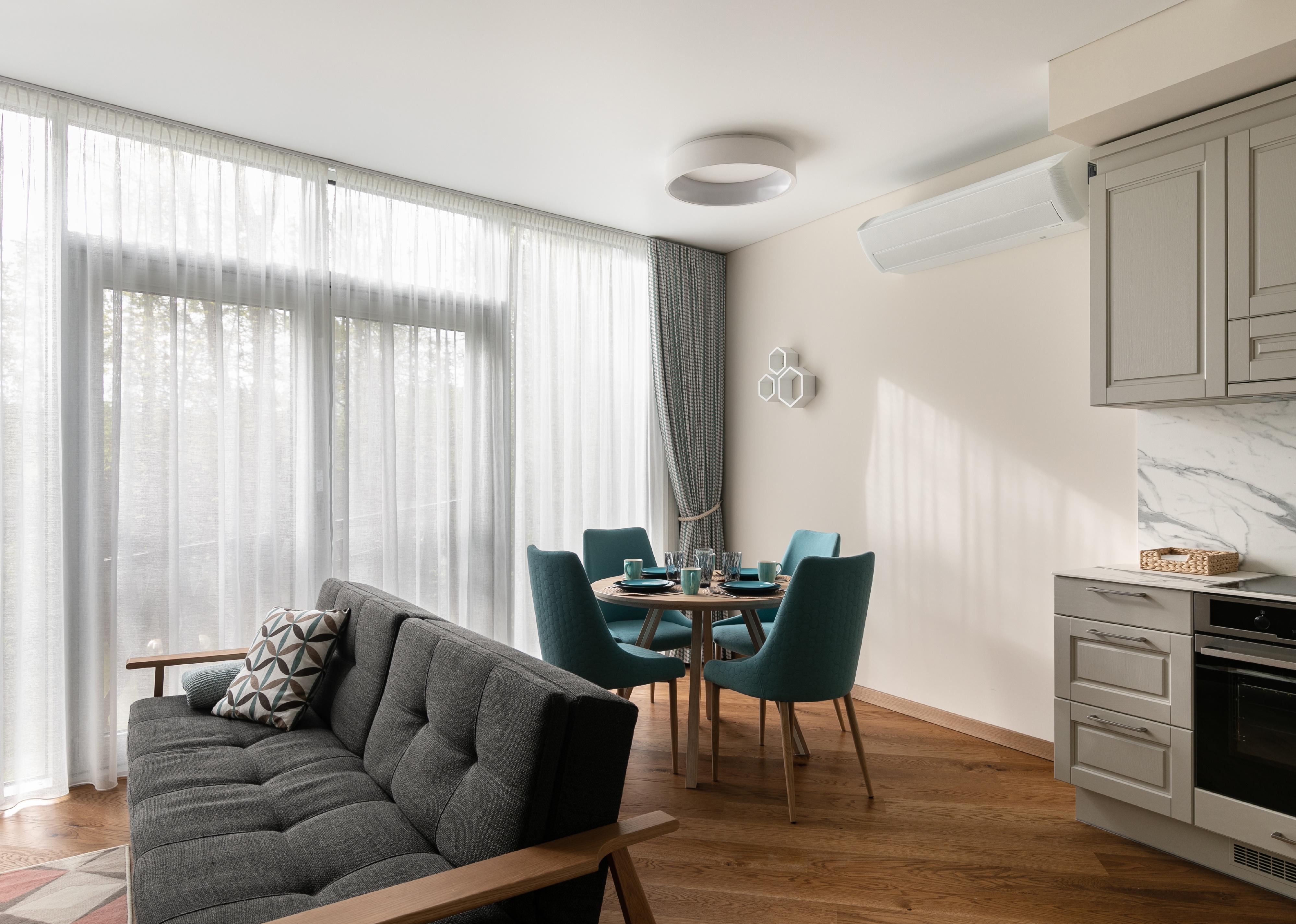 A photo of a living area with a grey sofa and dining setting with teal-green chairs, in front of a large window