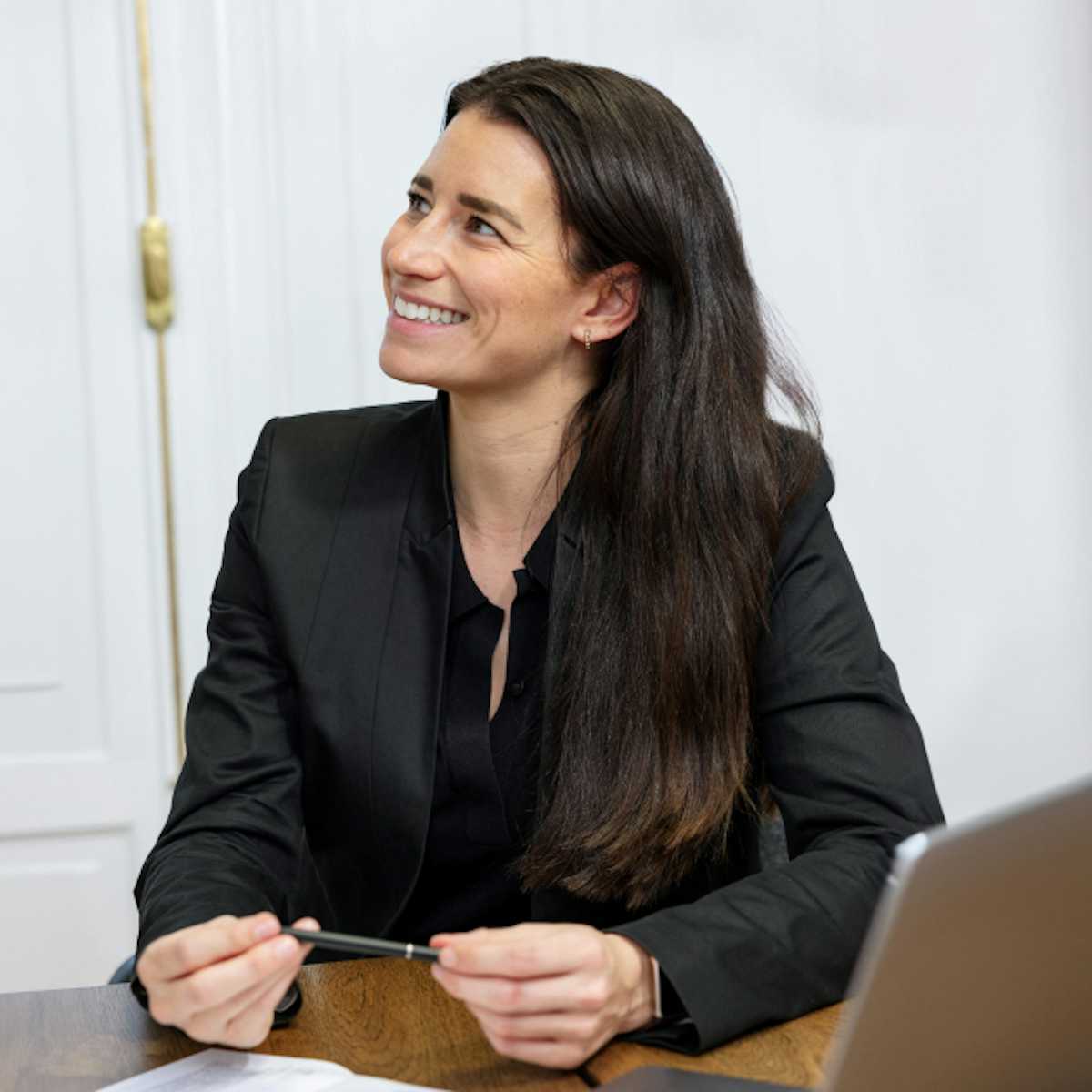 A woman sitting at a desk. There is a laptop on the table. She is holding a pen.