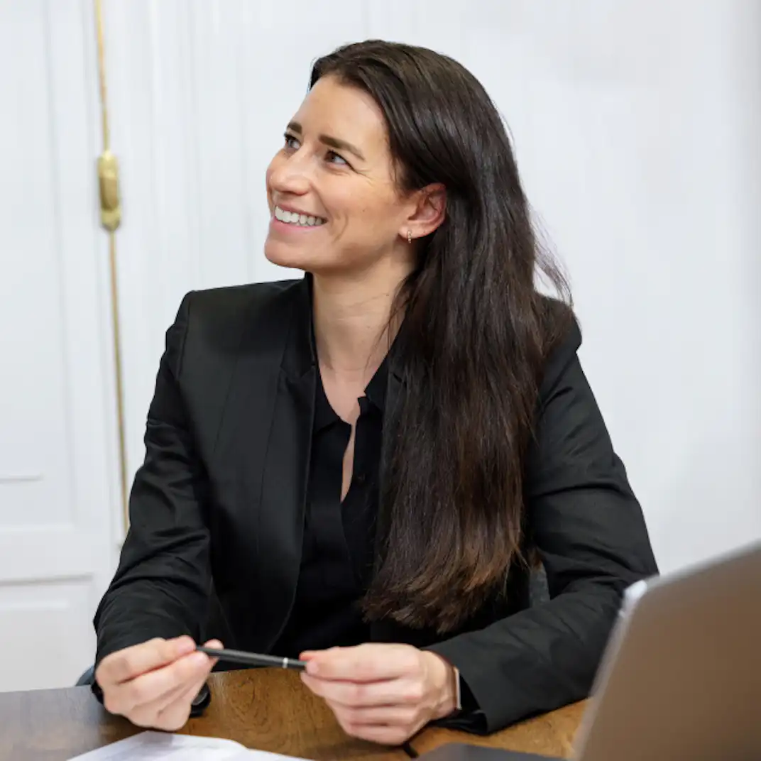 A woman sitting at a desk. There is a laptop on the table. She is holding a pen.