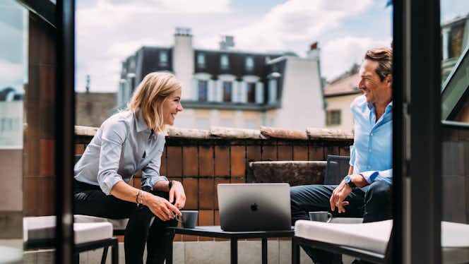 Two people sit on a balcony, smiling at each other over a laptop and coffee, with city rooftops in the background.