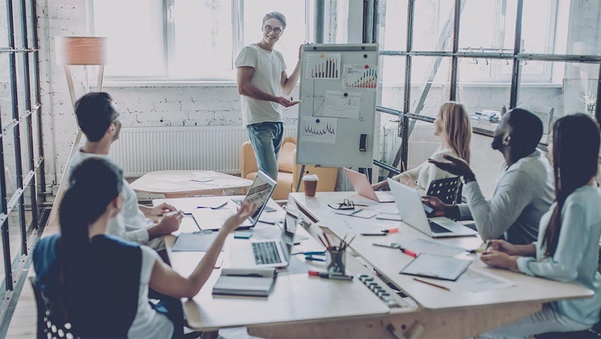 Équipe de personnes à une table de conférence avec quelqu'un qui présente sur un tableau blanc.