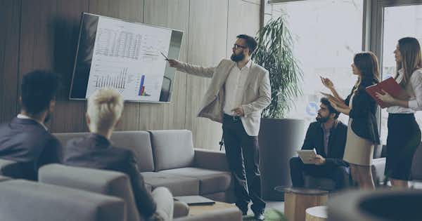 Man pointing to numbers on a large display in front of a group
